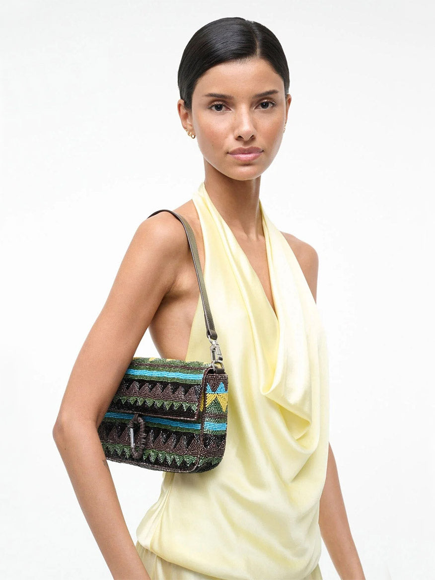Woman in a sleeveless pale yellow top carries the STAUD Timmy Shoulder Bag in Happy Sack, featuring geometric beaded designs and an interior zip pocket, standing against a plain white background.