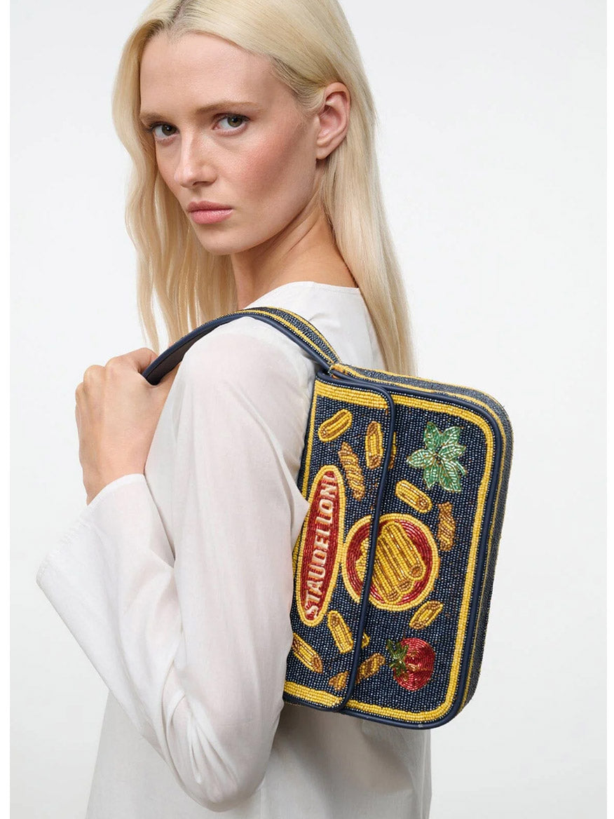 A woman with long blonde hair carries the limited-edition STAUD Tommy Beaded Bag in Staudelloni, featuring a hand-beaded burger, fries, and coins design on the side.