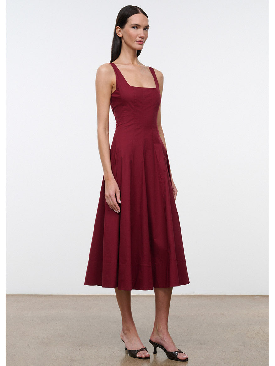 A woman stands indoors against a plain background, wearing the STAUD Wells Sleeveless Dress in Carmino—a burgundy cotton poplin dress with a square neckline—paired with black heeled sandals.