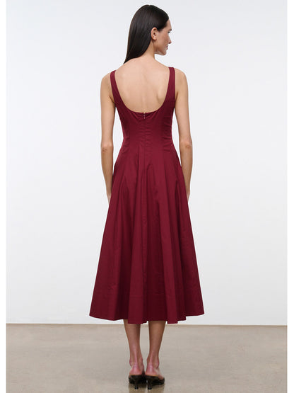 A woman stands facing away, wearing the STAUD Wells Sleeveless Dress in Carmino—a dark red cotton poplin dress with a fitted waist and flared skirt—set against a plain background.