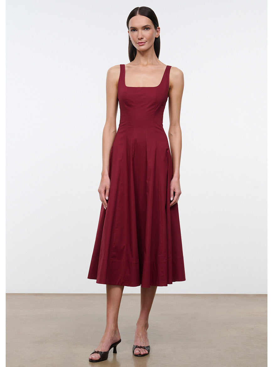 A woman models the STAUD Wells Sleeveless Dress in Carmino, featuring a square neckline and midi length in a rich burgundy shade, paired with black heeled sandals against a plain white background.