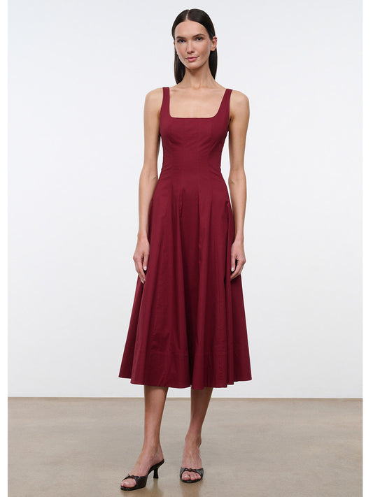 A woman models the STAUD Wells Sleeveless Dress in Carmino, featuring a square neckline and midi length in a rich burgundy shade, paired with black heeled sandals against a plain white background.