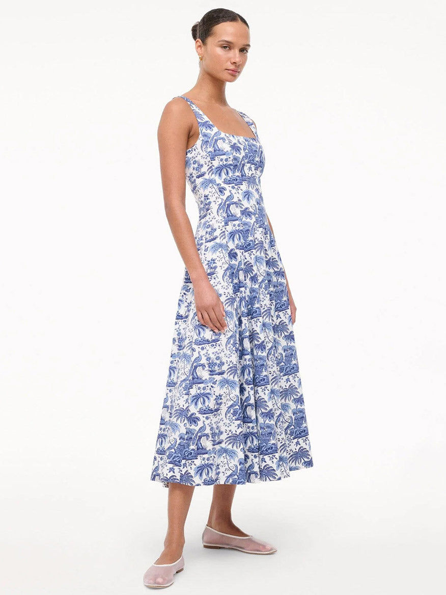 A woman wearing the STAUD Wells Sleeveless Dress in China Blue Toile with a fitted bodice and flared skirt, paired with light pink flat shoes, stands against a plain white background.