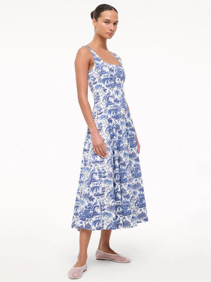 A woman wearing the STAUD Wells Sleeveless Dress in China Blue Toile with a fitted bodice and flared skirt, paired with light pink flat shoes, stands against a plain white background.