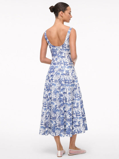 A woman stands facing away, wearing the STAUD Wells Sleeveless Dress in China Blue Toile—a blue and white cotton poplin, ankle-length dress with a floral pattern, scoop back, fitted waist. Her hair is tied back and she wears light pink flats.
