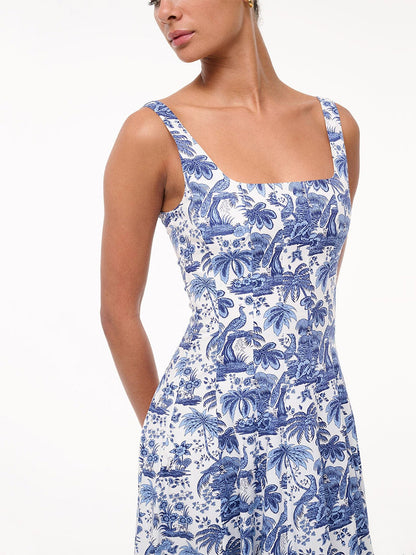 A woman wears the STAUD Wells Sleeveless Dress in China Blue Toile, featuring a square neck, fitted waist, and blue and white botanical and animal print, standing against a plain white background.