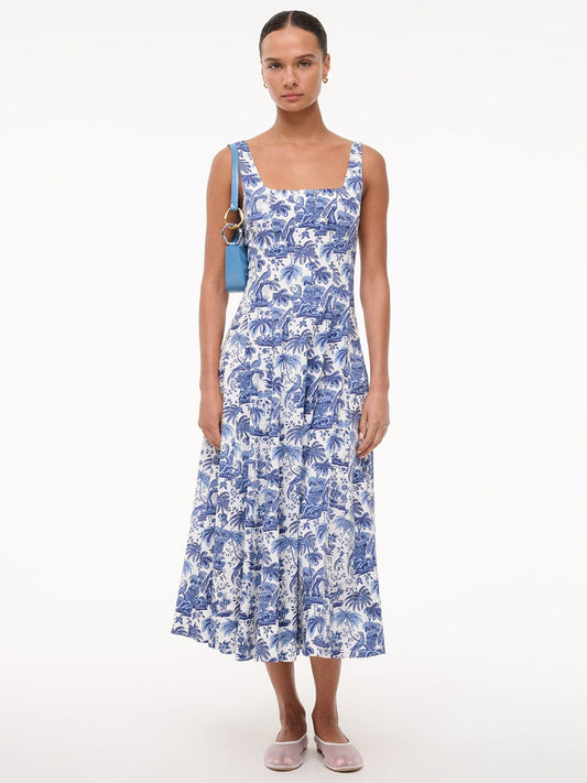 A woman stands against a white background wearing the STAUD Wells Sleeveless Dress in China Blue Toile, carrying a small blue bag and wearing flat pink shoes.