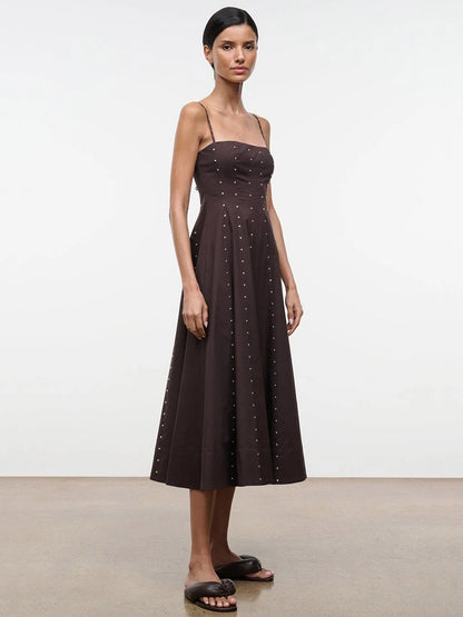 A woman wears the STAUD Wylie Dress in Earth Studs— a sleeveless, corset-style midi with spaghetti straps and silver studs—paired with black slide sandals, standing against a plain backdrop.