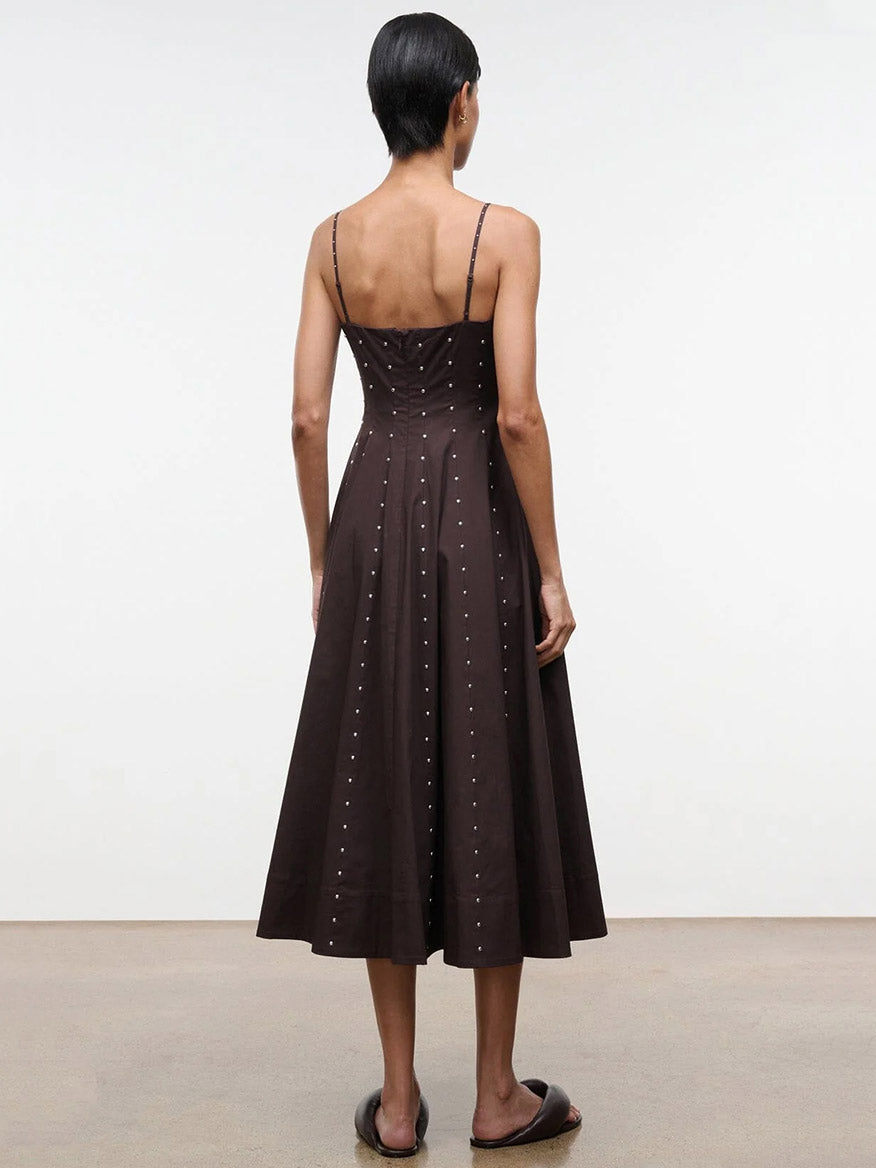 Someone stands indoors facing away, wearing the STAUD Wylie Dress in Earth Studs—a dark brown, calf-length corset-style dress with silver studs and thin straps—paired with black flat sandals.