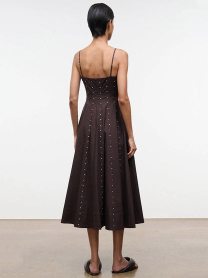 Someone stands indoors facing away, wearing the STAUD Wylie Dress in Earth Studs—a dark brown, calf-length corset-style dress with silver studs and thin straps—paired with black flat sandals.
