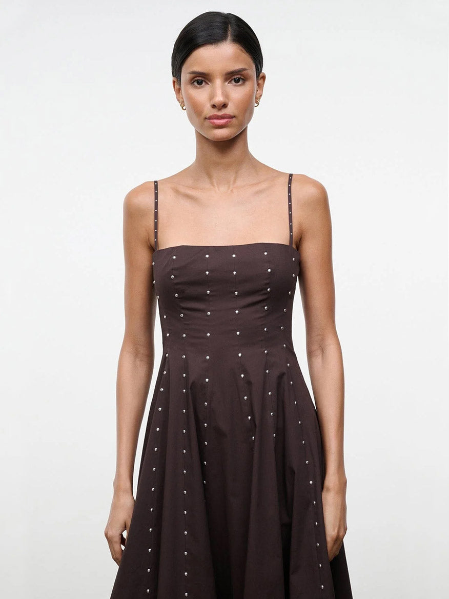 A woman with dark hair in a low bun models the STAUD Wylie Dress in Earth Studs, a sleeveless brown dress with spaghetti straps and small white dots, against a plain light background.