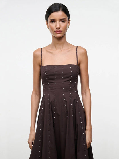 A woman with dark hair in a low bun models the STAUD Wylie Dress in Earth Studs, a sleeveless brown dress with spaghetti straps and small white dots, against a plain light background.
