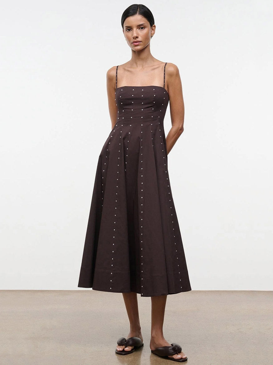 A woman stands against a plain background wearing the STAUD Wylie Dress in Earth Studs, a sleeveless, dark brown corset-style midi dress with thin straps and small button accents, paired with black flat sandals.