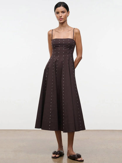 A woman stands against a plain background wearing the STAUD Wylie Dress in Earth Studs, a sleeveless, dark brown corset-style midi dress with thin straps and small button accents, paired with black flat sandals.