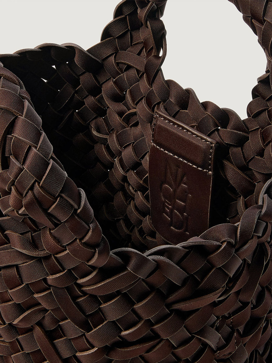 Close-up of a dark brown Naghedi St. Barths Petit Tote Nest Weave in Chocolate, a handwoven neoprene bag featuring an interior leather patch with an embossed logo.