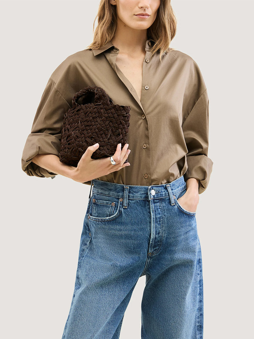 A person wearing a brown button-down shirt and blue jeans holds a small dark brown Naghedi St. Barths Petit Tote Nest Weave in Chocolate.