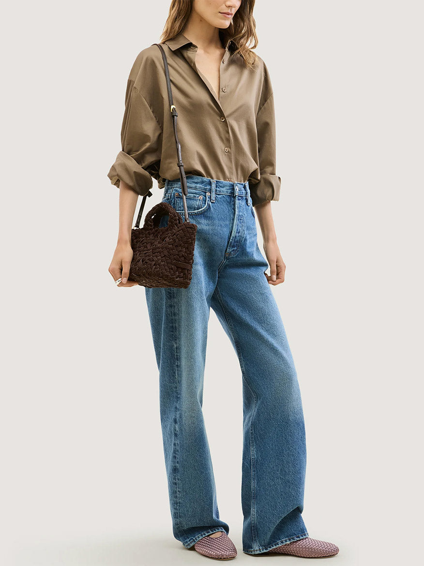 A person in a brown button-down shirt and blue jeans carries a small, dark brown handwoven Naghedi St. Barths Petit Tote Nest Weave in Chocolate as a crossbody purse.