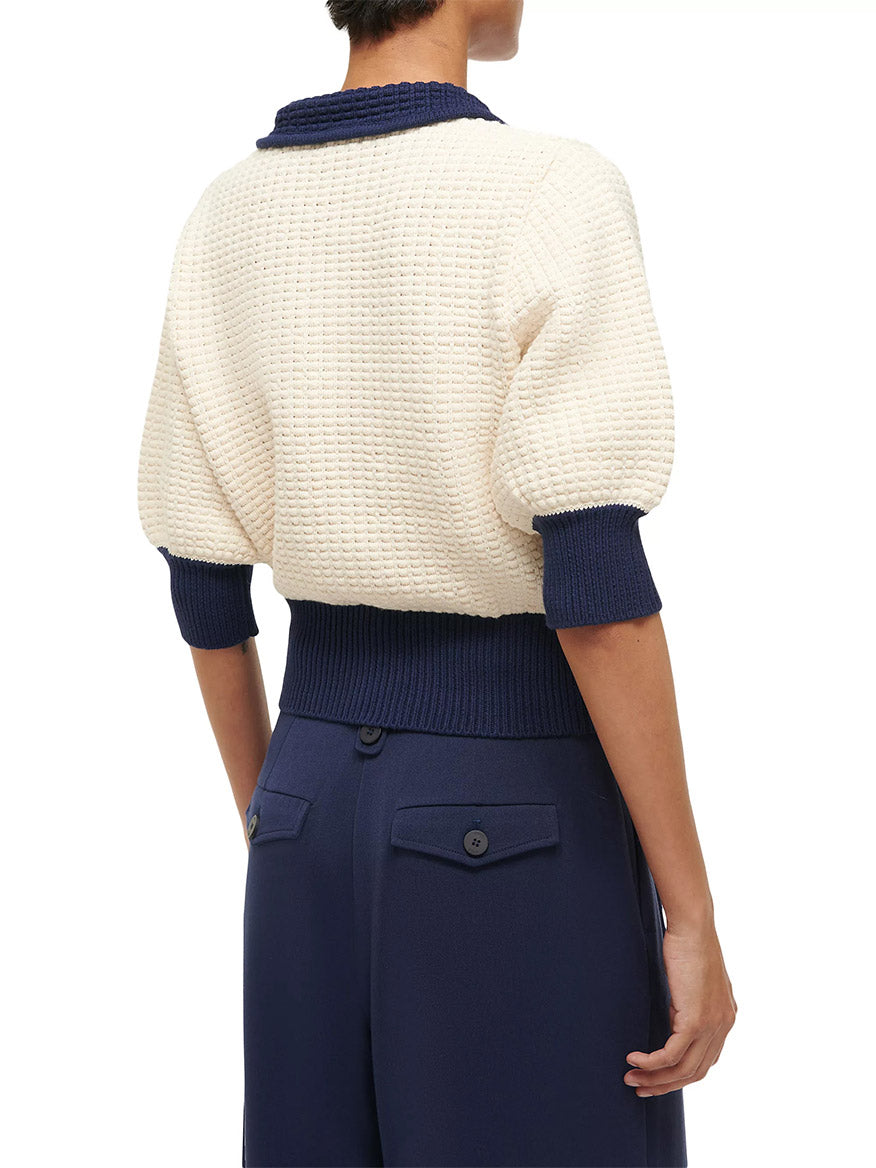 An individual dons the STAUD Altea Sweater in Ivory/Navy, featuring a cream textured design accented by navy blue cuffs and collar, perfectly paired with navy high-waisted pants. They stand with their back to the viewer, highlighting the sweater's chic cotton waffle knit.