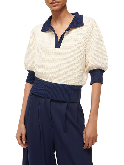 A person dressed in an ivory and navy STAUD Altea sweater, featuring a navy collar and cuffs, teamed with high-waisted navy pants.