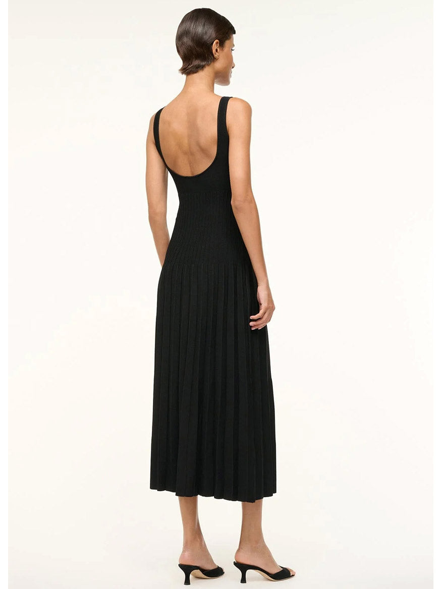 Dressed elegantly in a black STAUD Ellison Sleeveless Dress with ottoman stitch detailing and pleats, a person pairs it with sleek black heels while standing gracefully against a plain white background.