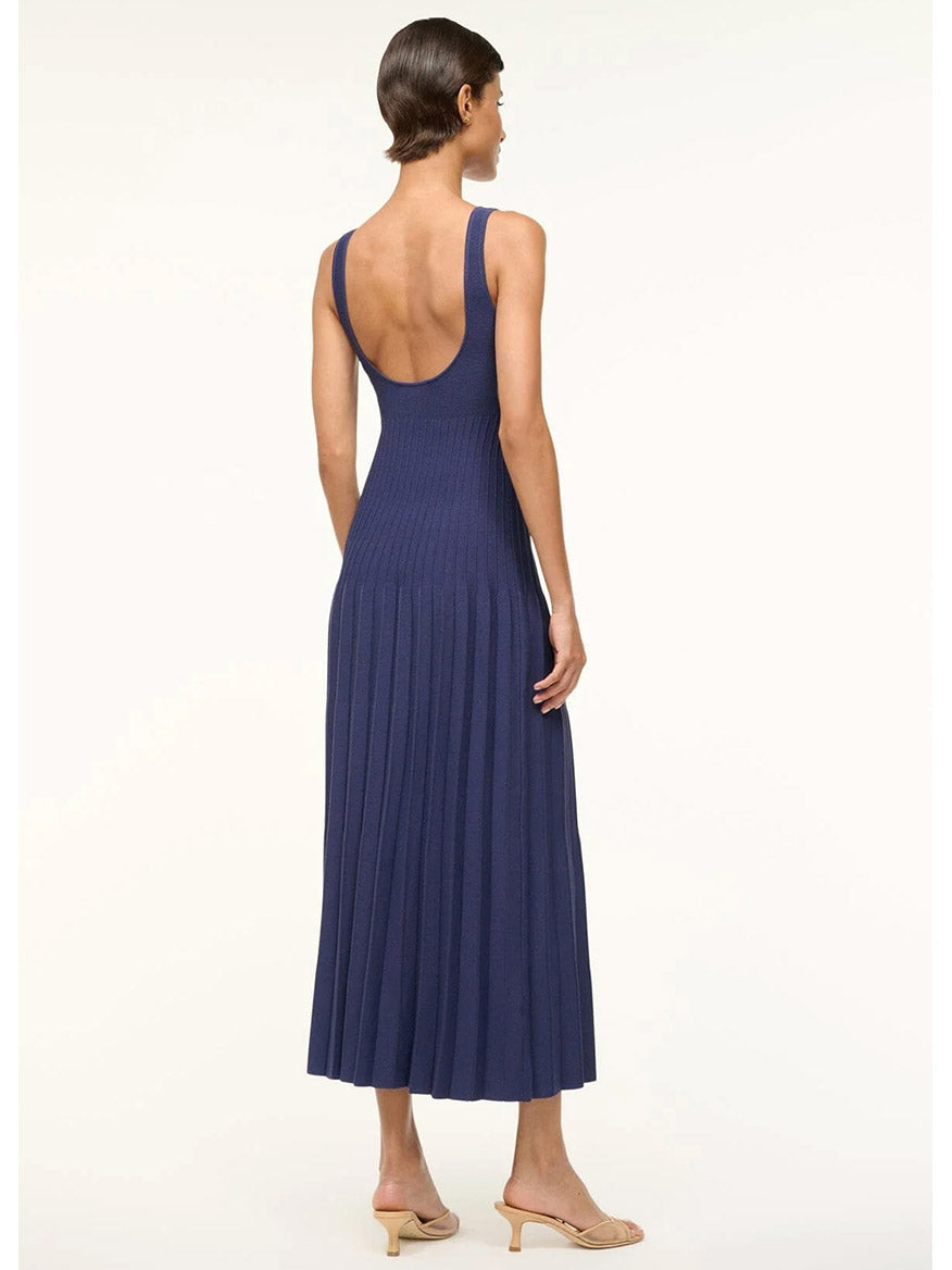 A person wearing the STAUD Ellison Sleeveless Dress in Navy, a backless design with ottoman stitch detailing and pleats, stands sideways against a white background, paired with beige heels.