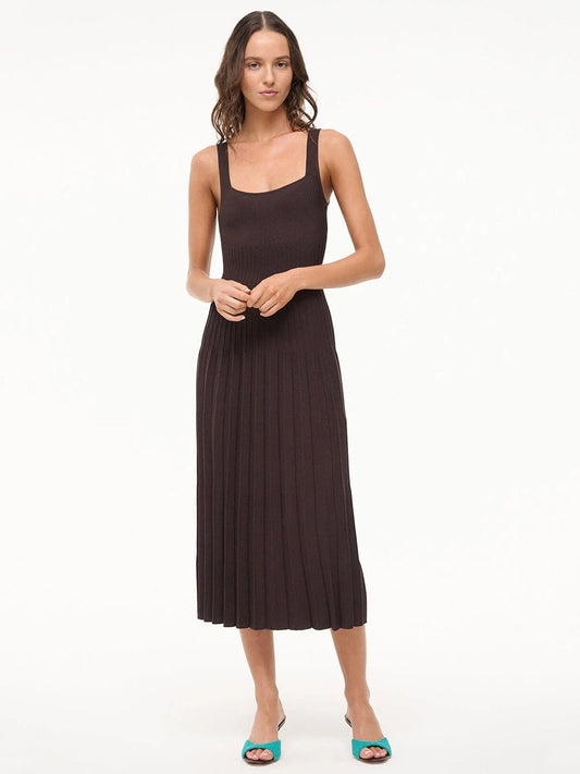 A woman stands against a plain background wearing the STAUD Ellison Sleeveless Dress in Earth—a dark brown, pleated midi style with a square neckline—paired with green open-toe heels.