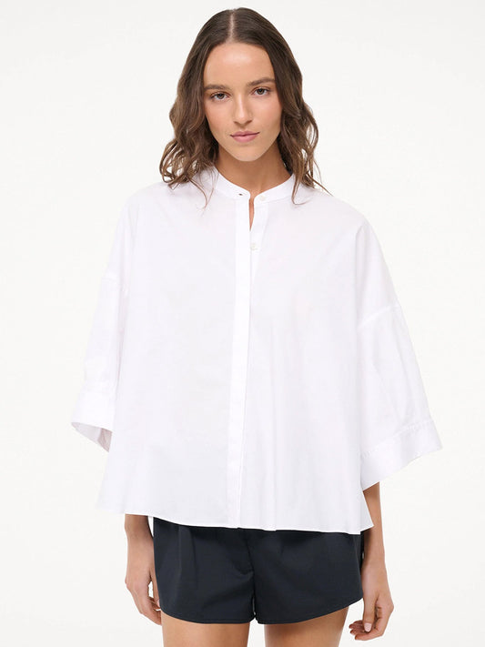 A woman stands facing forward against a plain light background, wearing the STAUD Enzo Top in white—a loose-fitting cotton poplin blouse with wide 3/4 length sleeves—paired with black shorts.