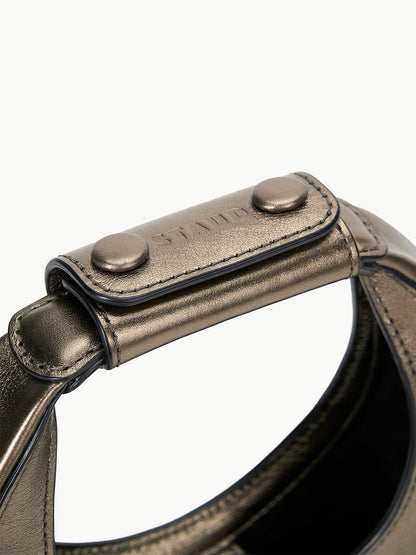 Close-up of the STAUD Goodnight Moon Bag in Aged Bronze showcasing a metallic handle with a stitched leather patch secured by two metal studs and complemented by a leather shoulder strap.