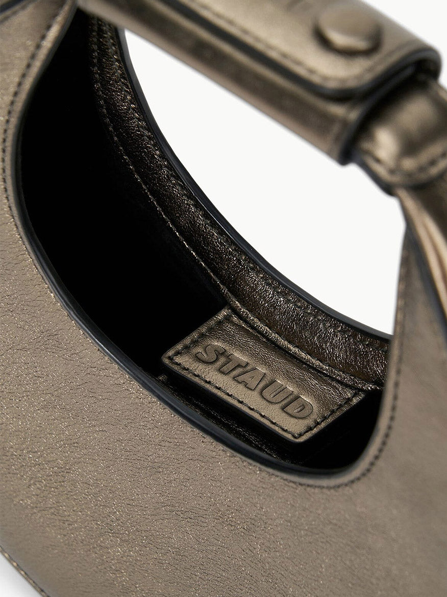 Close-up image of the STAUD Goodnight Moon Bag in Aged Bronze, a metallic leather evening bag with a shoulder strap, featuring an interior tag displaying the brand name "STAUD.