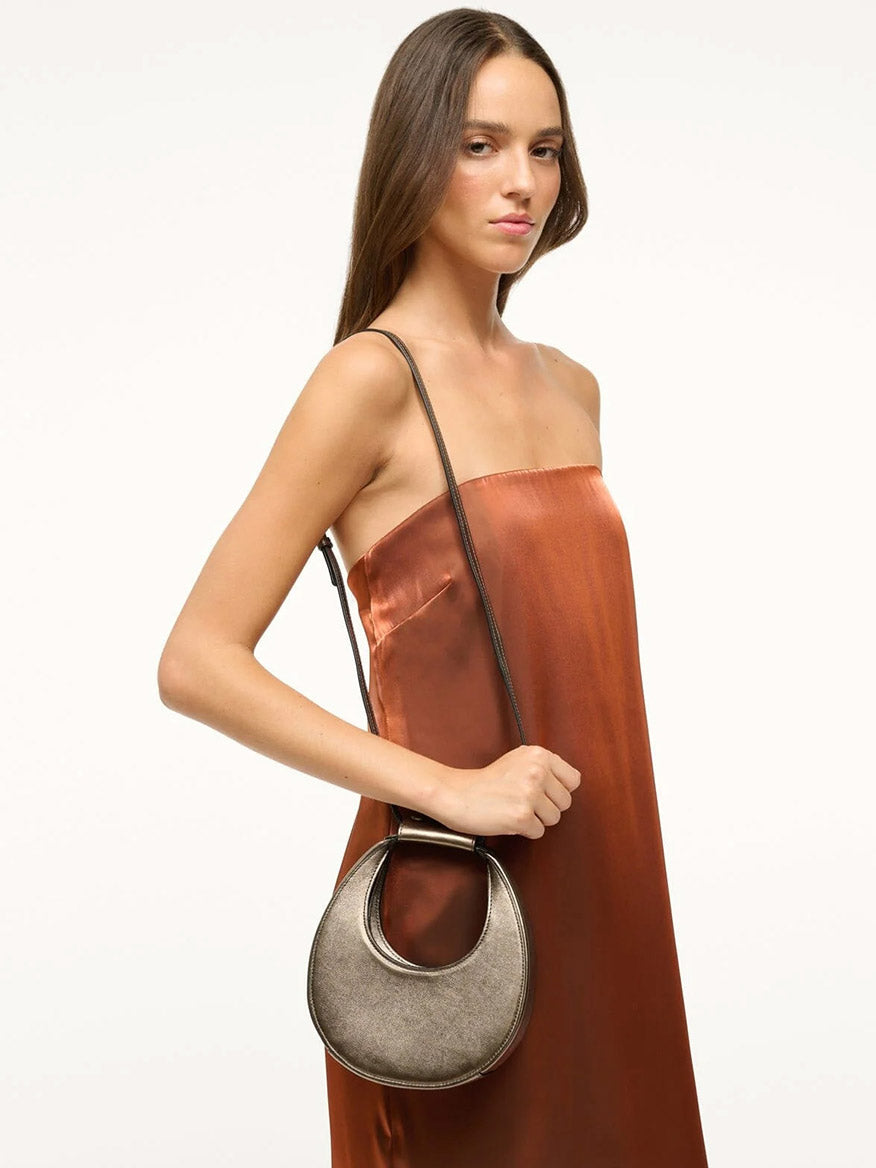 A woman in a strapless brown dress is holding the STAUD Goodnight Moon Bag in Aged Bronze, featuring a small size and a delicate leather shoulder strap.