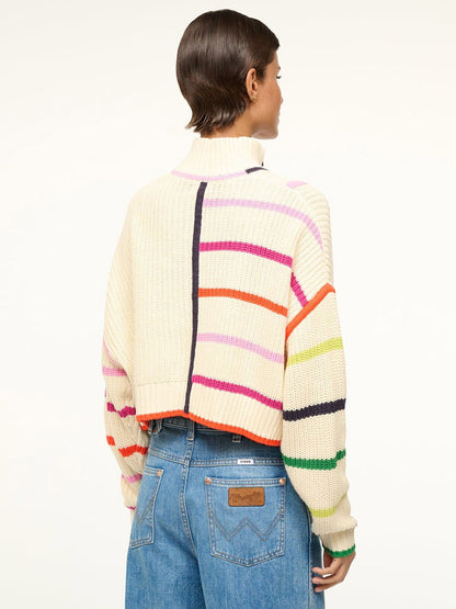 A person with short dark hair wears the STAUD Cropped Hampton Sweater in Cream/Rainbow Multi with blue jeans, its chunky knit and colorful stripes on display as they slightly turn away from the camera.