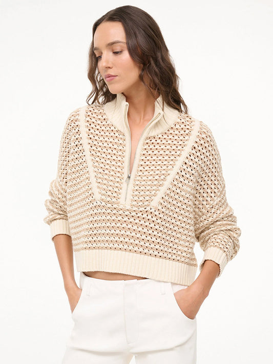 A woman wears the STAUD Cropped Hampton Sweater in Breeze Creme—a chunky cotton knit, cream half-zip—paired with white pants, standing with hands in pockets against a plain light background.