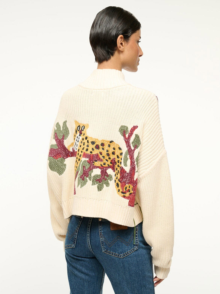 A person wearing the STAUD Cropped Hampton Sweater in Jungle Leopard, featuring a cream chunky cotton knit with a colorful jungle leopard and tree design on the back, paired with blue jeans.