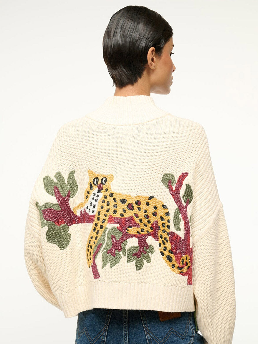 A person wearing the STAUD Cropped Hampton Sweater in Jungle Leopard, featuring a chunky cream cotton knit with a vibrant leopard and plant design on the back and a half-zip detail, looking to the side.