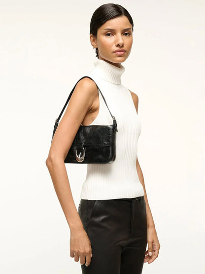 A woman in a sleeveless white turtleneck and black pants carries the STAUD Harlow Bag in Black over her shoulder.