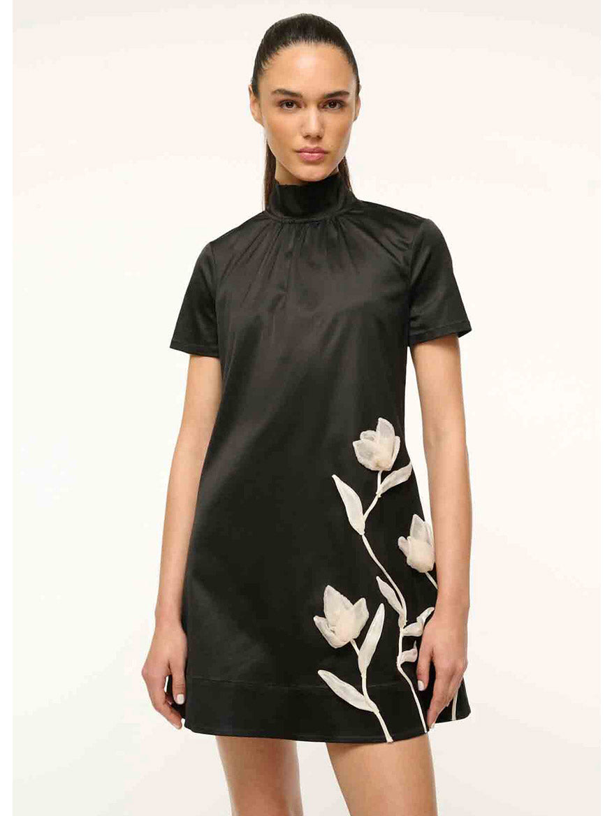 A person wearing the STAUD Mini Ilana Dress in black satin, featuring a high-neck and short sleeves with a white floral design, stands against a plain light background.