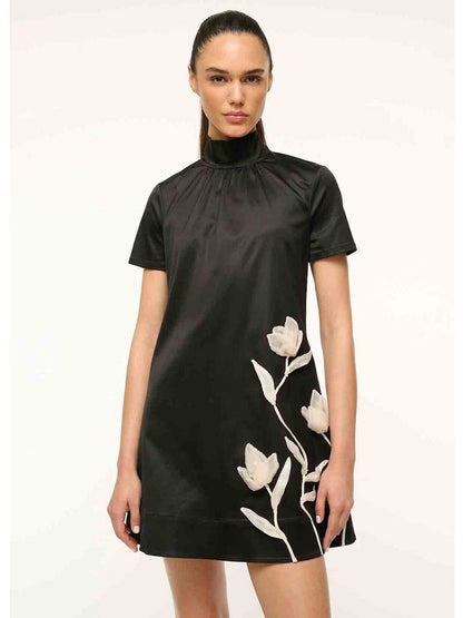 A person wearing the STAUD Mini Ilana Dress in black satin, featuring a high-neck and short sleeves with a white floral design, stands against a plain light background.