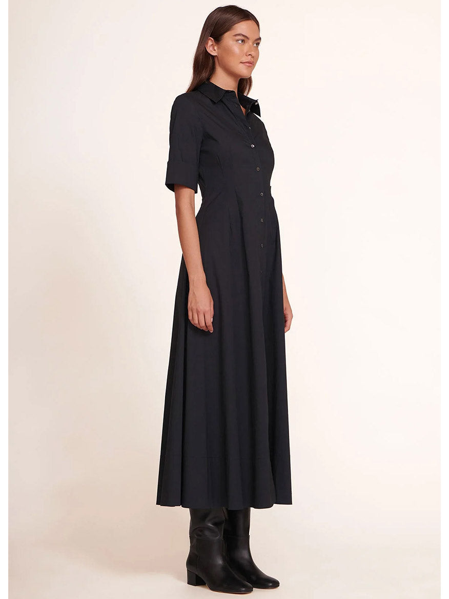 The woman stands in profile wearing the STAUD Joan Maxi Dress in black, a stretchy cotton poplin shirt dress with short sleeves. Paired with black boots against a plain background, her look embodies effortless elegance.