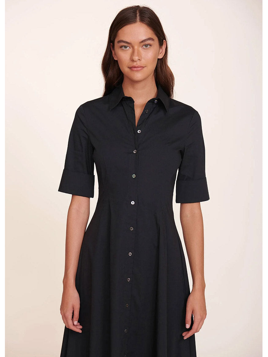 A woman models the STAUD Joan Maxi Dress in Black, a fitted maxi shirt dress crafted from stretchy cotton poplin, featuring short sleeves and a button-up design, as she stands facing forward against a plain background.