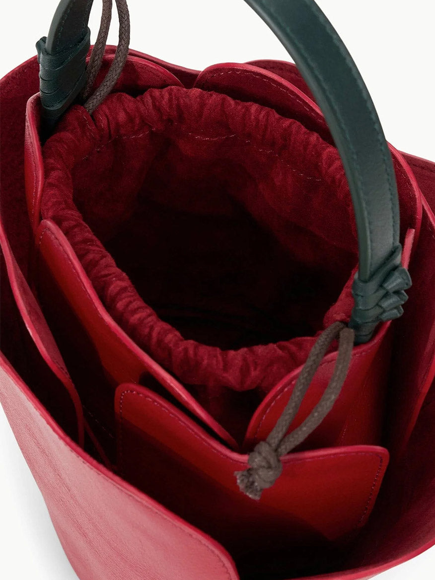 Presenting the STAUD Juliet Bag in Rouge—a captivating rose-shaped bucket bag crafted from beautiful red leather, complete with elegant dark green handles. This handheld accessory showcases an open compartment and a stylish red drawstring fabric interior, making it an ideal novelty piece to elevate any fashion-forward ensemble.