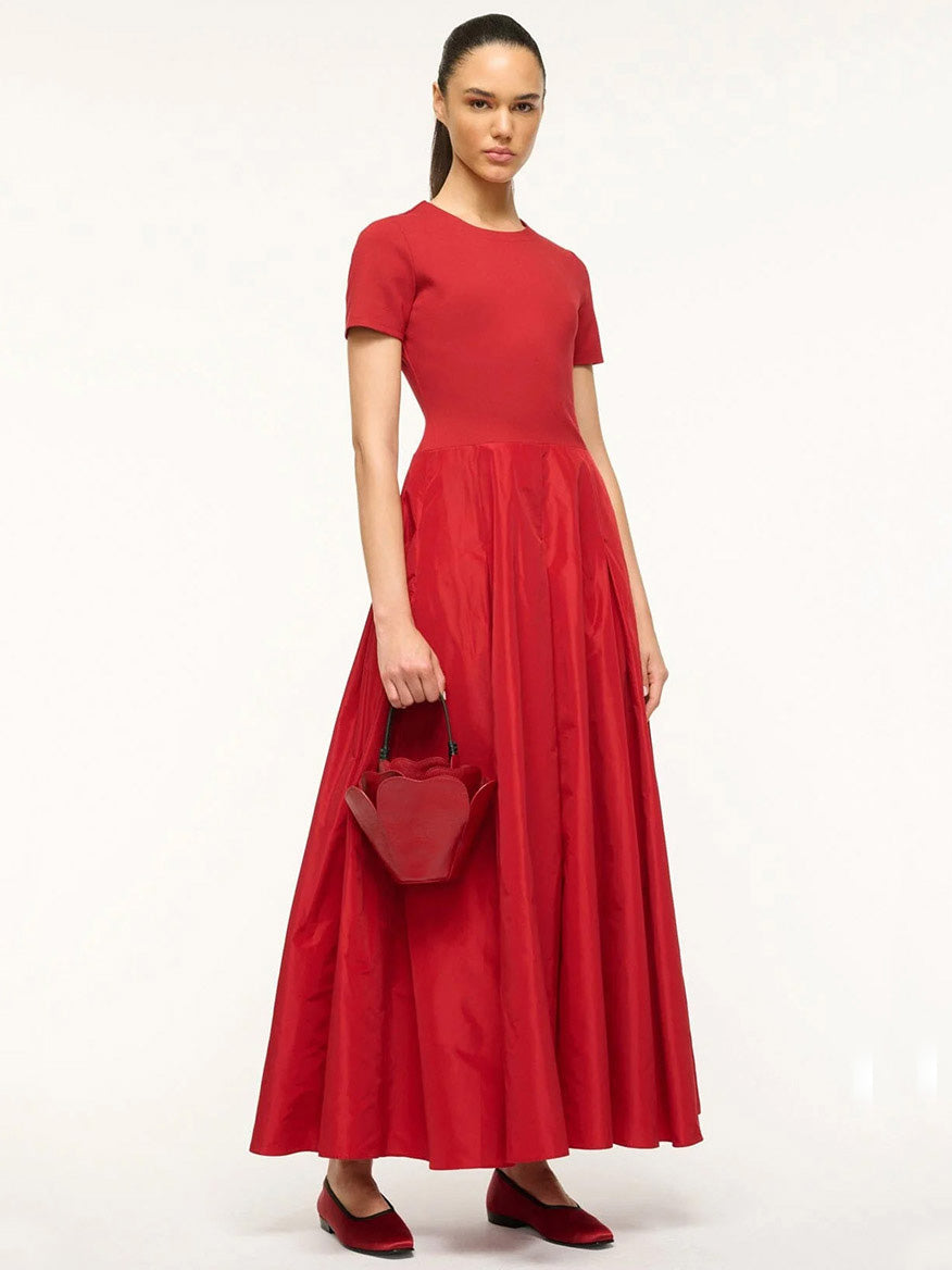 A woman stands in a red, short-sleeve, floor-length dress holding the STAUD Juliet Bag in Rouge, adding a touch of novelty to her ensemble.