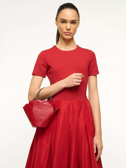 Dressed in red, a person holds the captivating STAUD Juliet Bag in Rouge, a striking novelty accessory against a plain background.