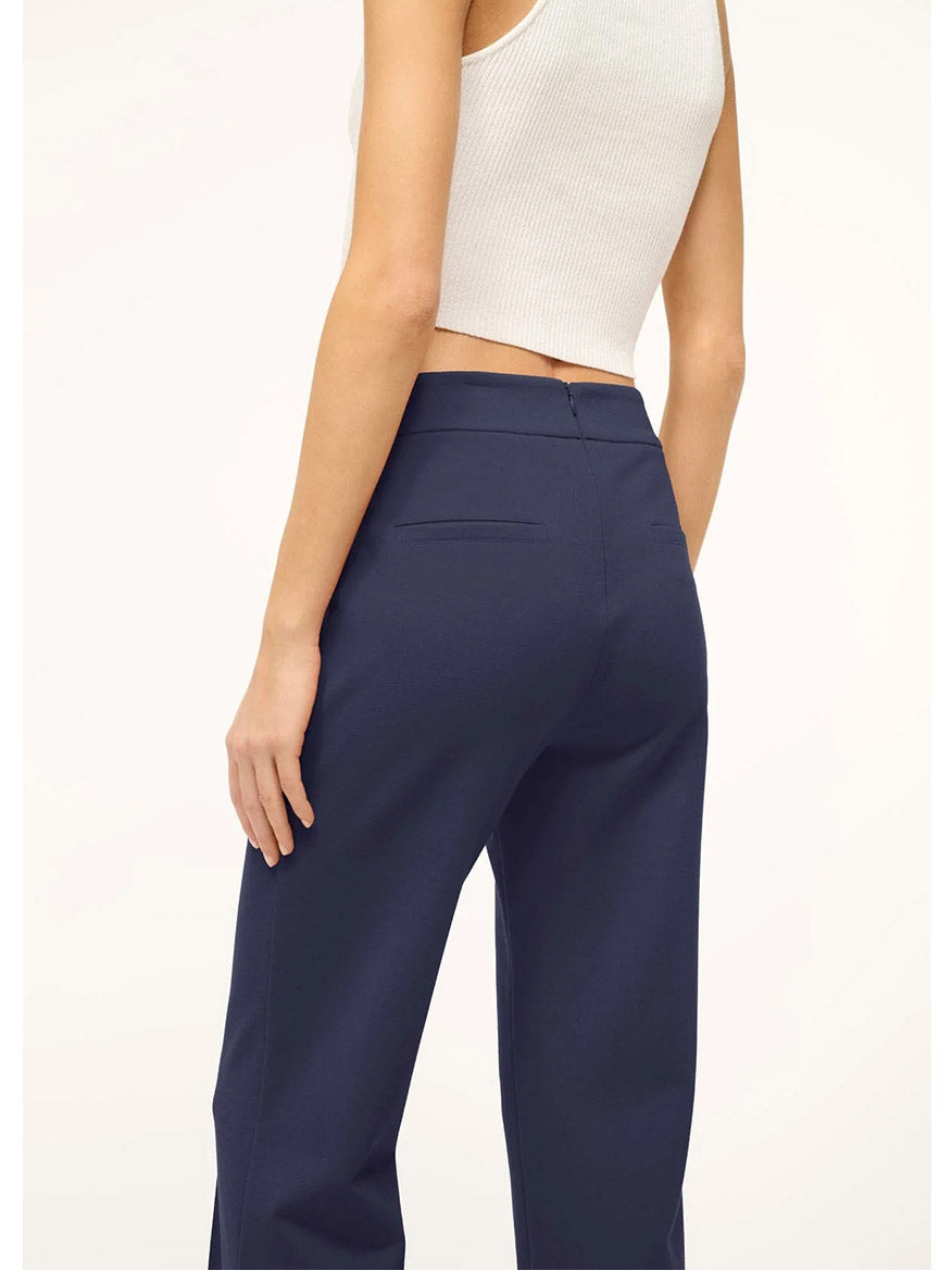 Someone is seen from the back, dressed in a matching set with a white sleeveless top and STAUD Knack Pant in Navy. The plain background accentuates the sleek lines of the stretchy ponte knit trousers.