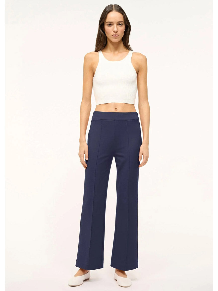 A person stands against a plain background wearing the STAUD Knack Pant in Navy, paired with a white sleeveless crop top and white shoes.