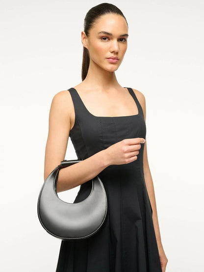 A woman in a black sleeveless dress holds the STAUD Mini Moon Bag in Black—a chic leather crescent bag—against a plain, light background.