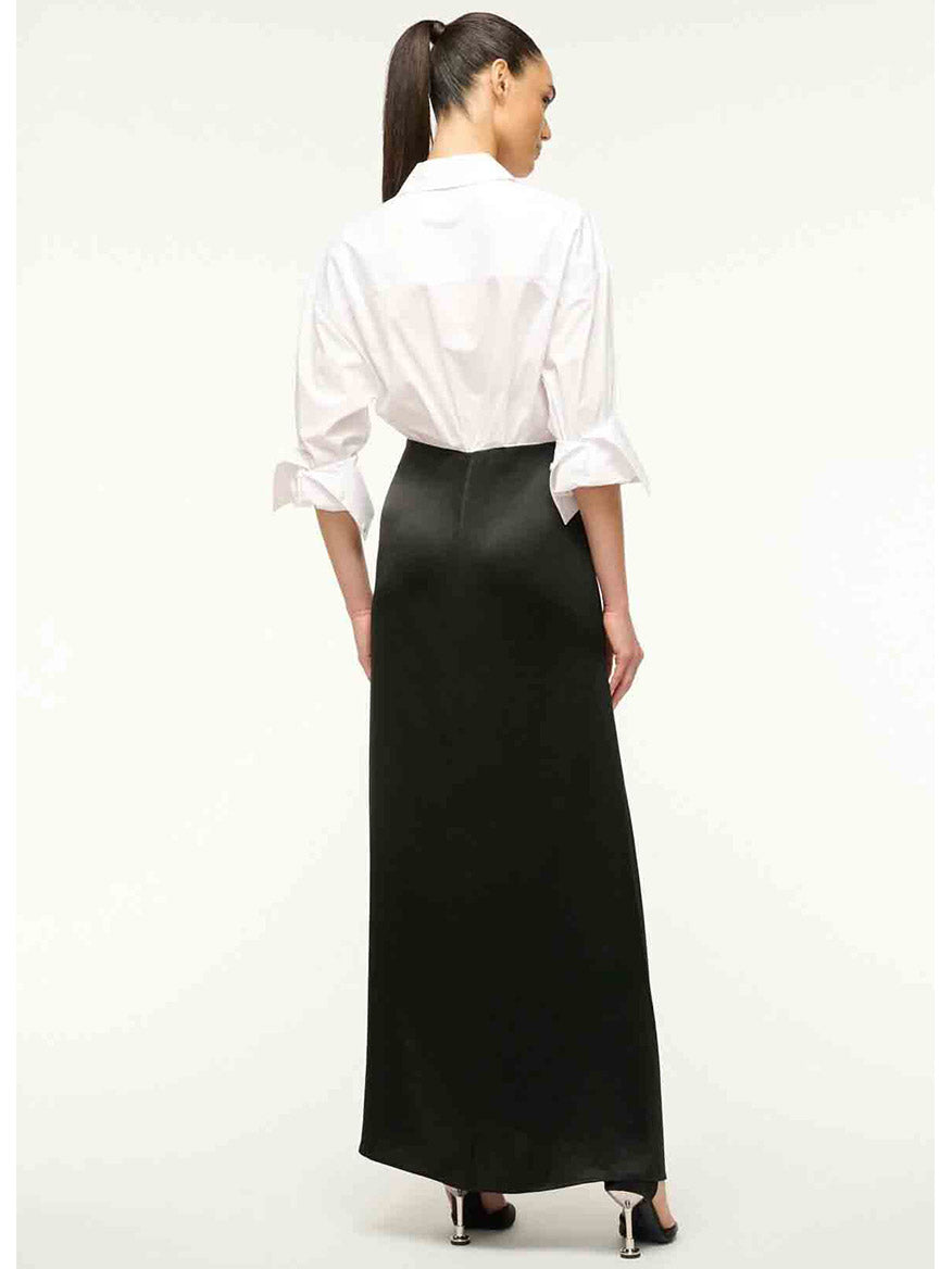 A woman is standing with her back to the camera, wearing the STAUD Nile Dress in Black, featuring a white poplin shirt bodice and a long black satin skirt. Her hair is styled in a ponytail.