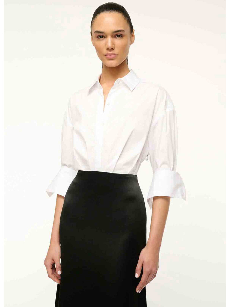 A person in the STAUD Nile Dress, featuring a white poplin shirt bodice with rolled sleeves and a sleek black satin skirt, stands against a plain background.