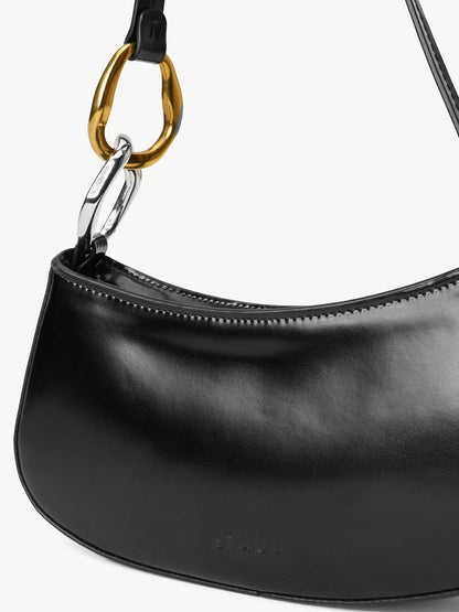 A close-up of the STAUD Ollie Bag in Black, a polished leather mini shoulder bag with gold and silver two-tone hardware ring accents near the strap attachment.