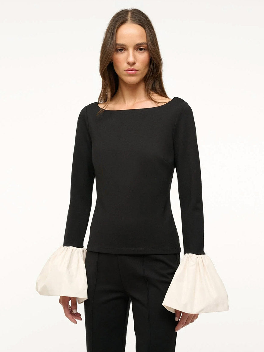 A woman stands against a plain background wearing the chic STAUD Paige Contrast Sleeve Top in Black/Ivory, featuring elegant organza cuffs and paired with comfortable black ponte fabric pants. She has long hair and a neutral expression.