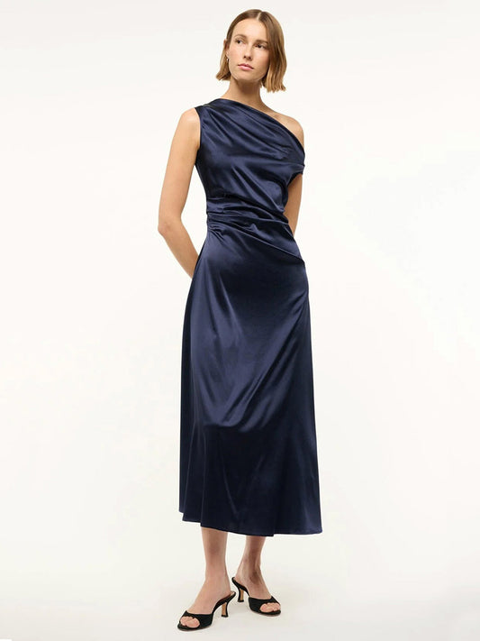 A woman wears the STAUD Phare Silk Dress in Navy—a shiny, stretch silk one-shoulder dress with waist ruching—paired with black heeled sandals against a plain light background, perfect for special occasions.